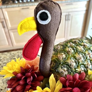 May include: A Thanksgiving centerpiece featuring a brown felt turkey with a yellow beak and red wattle, perched atop a pineapple surrounded by red, yellow, and orange flowers.
