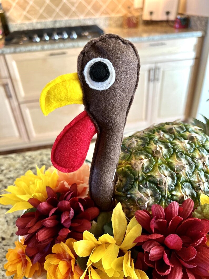 May include: A Thanksgiving centerpiece featuring a brown felt turkey with a yellow beak and red wattle, perched atop a pineapple surrounded by red, yellow, and orange flowers.