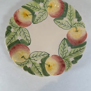 May include: A round, off-white ceramic plate with a hand-painted apple and leaf design. The rim features a repeating pattern of red and yellow apples, green leaves, and small white flowers. The plate has a scalloped edge.