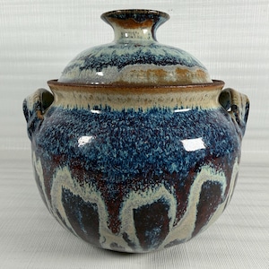 May include: A handcrafted ceramic casserole dish with a lid. The dish features a blue, brown, and cream glaze. It has two handles and a knob on the lid. The design has a rustic, artisanal aesthetic.