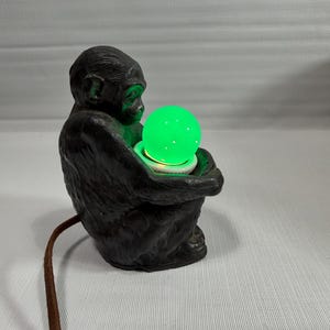 May include: A dark brown, textured monkey-shaped lamp holding a glowing, spherical green light bulb. The lamp features a detailed, sculpted primate figure in a seated position, with a brown electrical cord extending from the base.