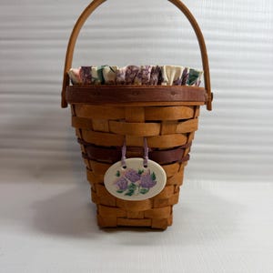 May include: A woven basket with a wooden handle and a floral fabric lining. The basket has a decorative oval tag with a purple flower design. The basket is made of light brown and dark brown woven material.