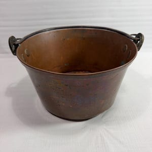 May include: A round copper pot with a dark, weathered patina and a wide, open top. The vessel features a sturdy, dark metal swing handle attached to two side loops, with visible rivets securing the handle mounts to the interior walls.