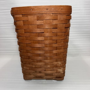 May include: A rectangular, woven basket in shades of brown. The basket is made of thin strips of wood, tightly woven together. The basket has a slightly tapered shape, wider at the top and narrower at the base. The basket is empty.