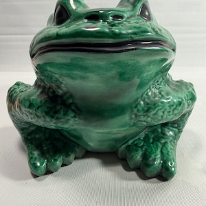 May include: A ceramic frog figurine featuring a glossy, deep green glaze with darker shaded accents. The frog is depicted in a seated, forward-facing position with a wide, smooth mouth, prominent eyes, and textured skin details on its limbs.