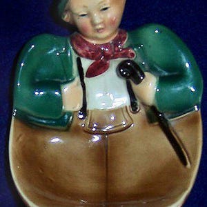 May include: A vintage ceramic figurine of a young boy in traditional attire. The figurine features a green jacket, brown pants, a white shirt, and a red scarf. The boy is holding a cane and a walking stick. The figurine is on a dark blue background.