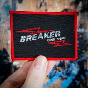 Könnte beinhalten: Schwarzer rechteckiger Aufnäher mit rotem Rand. Der Aufnäher zeigt den Text "BREAKER ONE NINE" in Silber und Rot, mit roten Blitzdesigns. Der Aufnäher wird von einer Hand gehalten.
