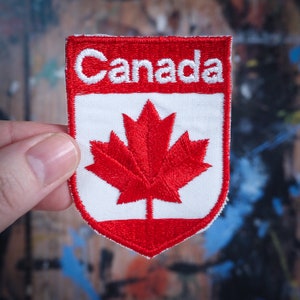 May include: Red, white and red embroidered patch with the word "Canada" and a red maple leaf on a white background.