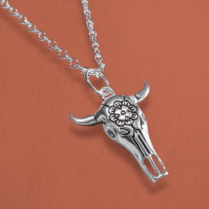 May include: A silver-toned necklace featuring a bull skull pendant. The pendant has a detailed floral design on the forehead and curved horns. The chain is a delicate link style, and the background is a solid orange color.