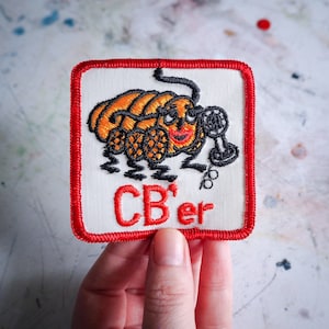 Könnte beinhalten: Ein roter, weißer und schwarzer gestickter Aufnäher mit einem Cartoon-Käfer, der ein Mikrofon hält. Der Text "CB'er" ist in Rot auf dem Aufnäher gestickt.