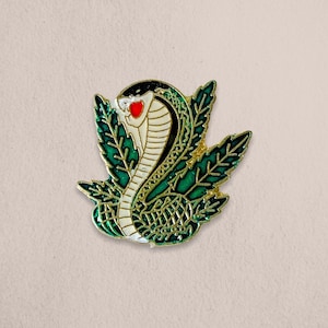 May include: A gold-colored enamel pin featuring a green and white cobra with a red heart, surrounded by green leaves.