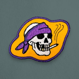 May include: Purple and white embroidered patch of a skull wearing a purple bandana and smoking a cigarette. The skull is on a yellow background with a purple outline.