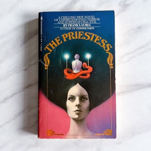 May include: A blue book cover with the title "The Priestess" in gold lettering. The cover features a woman with dark hair and a red snake around her head. The book is a novel by Frank Lauria, author of Communion.
