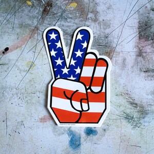 May include: A peace sign sticker with the American flag design. The sticker is red, white, and blue.