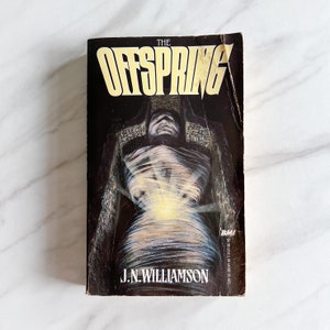 May include: A paperback book cover with a dark background and a white figure in the centre. The title "THE OFFSPRING" is printed in yellow letters at the top of the cover. The author's name, "J.N. WILLIAMSON", is printed in white letters at the bottom of the cover.
