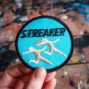 May include: A blue and black embroidered patch with the word "Streaker" in black lettering and two white figures running on a blue background.