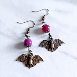 May include: A pair of dangle earrings featuring a small bat charm and a dark pink round bead. The earrings are made of metal and have a bronze finish.