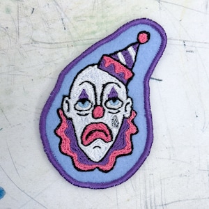 May include: An embroidered patch featuring a sad clown with a pink and white hat. The clown has a white face with pink lips, a red nose, and purple eyebrows. The patch is purple and blue.