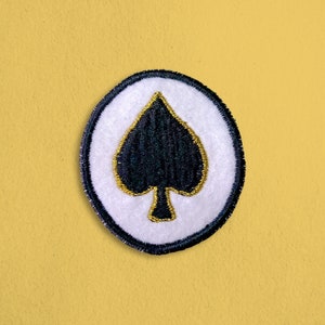 May include: A white circle with a black spade in the center. The spade is outlined in gold thread. The circle is surrounded by a black border.