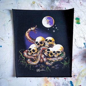 May include: Black patch with a colorful image of a snake wrapped around a pile of skulls. The snake is red, yellow, and black. The skulls are white and have black eye sockets. There is a purple moon in the background with stars. The patch has the text "ZIP LONDON ©" in the bottom right corner.