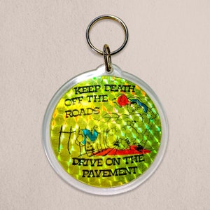 Könnte beinhalten: Ein schillernder Schlüsselanhänger in Gelb und Grün mit dem Text "Keep Death Off The Roads Drive On The Pavement". Der Schlüsselanhänger zeigt ein Cartoon-Bild einer Person, die ein Auto fährt.