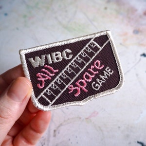 May include: A brown embroidered patch with white stitching. The patch features the text "WIBC All Spare Game" in white and pink lettering.