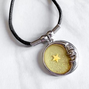 Könnte beinhalten: Eine silberne Mondsichel-Anhängerkette mit einem gelben Stern im Inneren. Der Anhänger ist mit Glitzer gefüllt und hat eine schwarze Schnur.