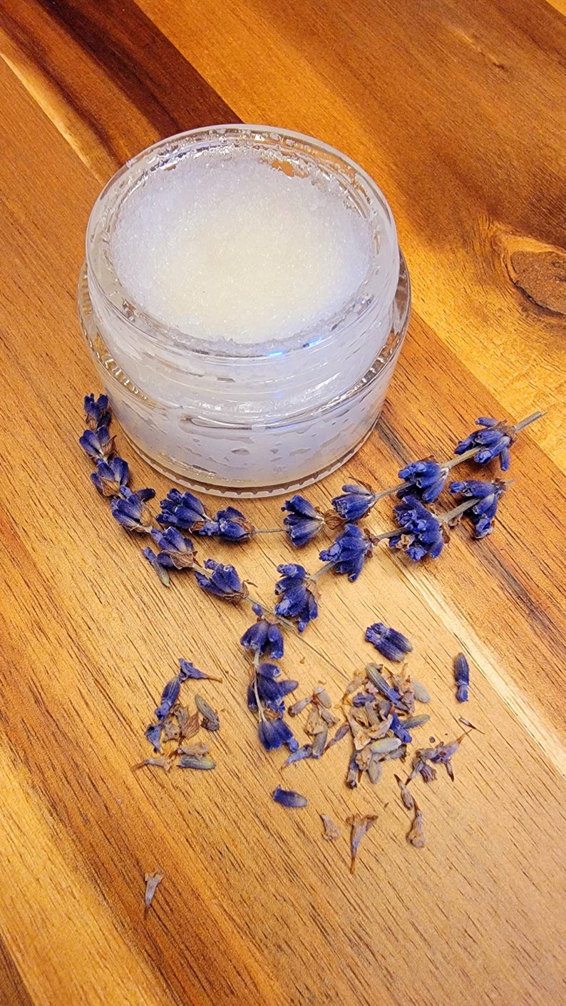 Puede incluir: Un peque&ntilde;o frasco de vidrio lleno de una sustancia blanca y granulada, posiblemente un exfoliante de az&uacute;car. El frasco est&aacute; rodeado de flores de lavanda secas sobre una superficie de madera.