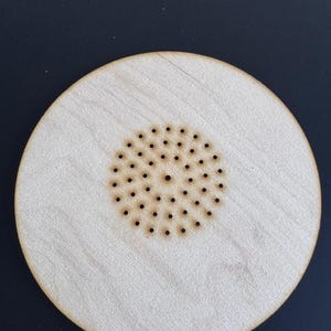 Op de afbeelding: Een ronde, lichtgekleurde houten onderzetter met een centraal patroon van kleine, gelijkmatig verdeelde gaatjes. De onderzetter heeft een natuurlijke houtnerf en een eenvoudig, minimalistisch ontwerp. De onderzetter heeft een diameter van ongeveer 10 cm.