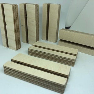 May include: Set of six wooden display stands with a single slot for holding cards or photos. The stands are made of light brown plywood and have a natural finish.