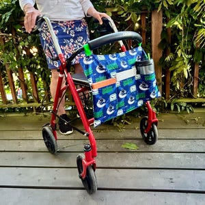 Peut inclure: Déambulateur rouge avec des roues noires et un sac bleu et blanc à motifs avec le logo des Canucks de Vancouver. Le sac comporte des poches et un porte-bouteille. Le déambulateur est sur une terrasse en bois.