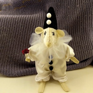 Könnte beinhalten: Handgefertigte Mäusefigur in einem Pierrot-Kostüm, mit einem weißen Satin-Outfit, Rüschenkragen und einem schwarzen Hut mit weißen Bommeln. Die Maus hält eine kleine rote Rose. Der Hintergrund zeigt einen grauen Strickstoff.