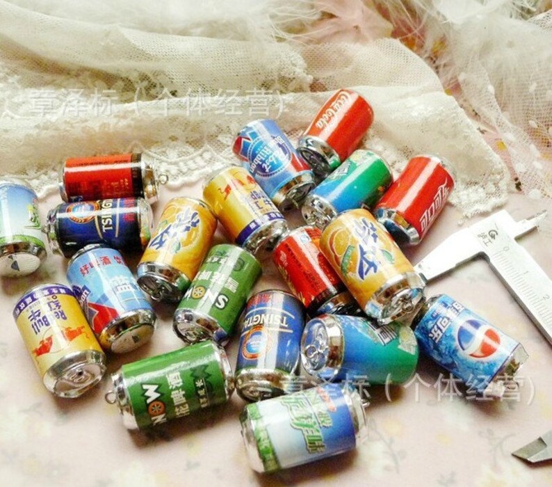 May include: A collection of miniature aluminum beverage cans with various brand logos and designs. The cans are silver with colorful labels and are arranged on a white surface.