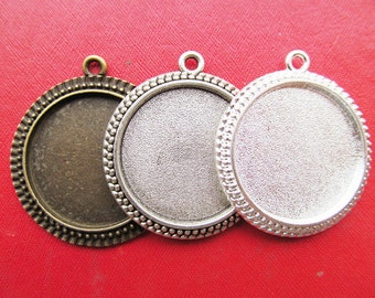 May include: Three round metal pendant trays in antique bronze, silver, and pewter. Each has a textured interior and a decorative beaded edge, with a small loop at the top for attaching to a chain or cord. The trays are displayed on a red surface.