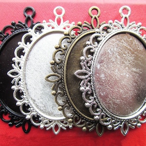 Puede incluir: Cuatro bandejas de colgante ornamentadas en negro, plata, bronce y plata antigua. Cada bandeja de colgante tiene un borde de filigrana decorativo y un engaste circular para un cabujón u otro inserto. Las bandejas se muestran sobre una superficie roja.