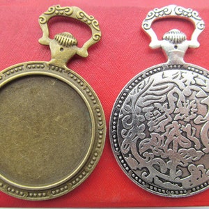 Pode incluir: Dois pendentes em branco de metal de estilo antigo com um design circular. O pendente à esquerda é de bronze com uma superfície lisa e uma fivela decorativa. O pendente à direita é de prata com um padrão floral intrincado e uma fivela decorativa.