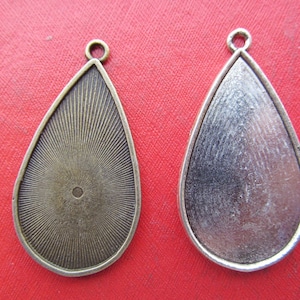 Könnte beinhalten: Zwei tropfenförmige Anhänger-Tabletts. Eines hat ein bronzefarbenes Sonnenstrahl-Design, das andere eine silberfarbene, gebürstete Metalloberfläche. Beide haben oben eine kleine Schlaufe zum Befestigen an einer Kette oder einem Band. Für die Schmuckherstellung.