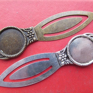 Puede incluir: Dos colgantes de marcapáginas de metal con un espacio redondo en blanco para agregar un amuleto o una foto. Un marcapáginas es plateado con un espacio redondo plateado y el otro es bronce con un espacio redondo bronce.