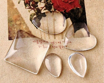 May include: Five clear glass cabochons of different shapes and sizes. The cabochons are on a brown surface with a vintage postcard in the background.