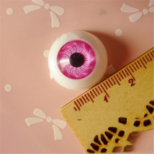 May include: A close-up of a pink and white plastic doll eye with a black pupil. The eye is approximately 2.5 cm in diameter and is sitting on a pink surface next to a ruler.