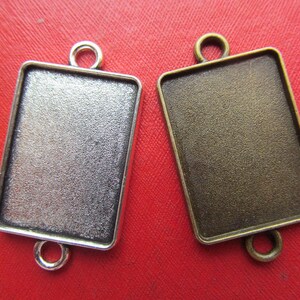 May include: Two rectangular metal picture frames with a silver and bronze finish. Each frame has a loop at the top for hanging and a bail at the bottom for attaching to a chain or cord.