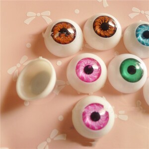 May include: Assortment of colourful plastic doll eyes with different iris patterns. The eyes are white with a black pupil and various coloured irises, including brown, green, pink, and blue. The eyes are displayed on a pink surface.