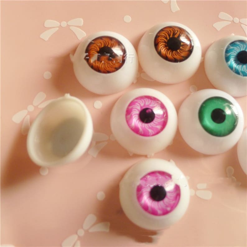May include: Assortment of colourful plastic doll eyes with different iris patterns. The eyes are white with a black pupil and various coloured irises, including brown, green, pink, and blue. The eyes are displayed on a pink surface.