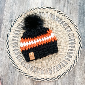 May include: Black, orange, and white crocheted beanie with a black pom pom. The beanie has a brown leather tag that says "Stitch Fiesta".