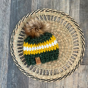 May include: Green, yellow, and white crocheted beanie with a brown faux fur pom pom. The beanie has a brown leather tag that says "Stitch Fiesta".