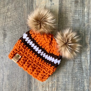 May include: Orange, brown, and white striped crocheted beanie with two brown faux fur pom poms. The beanie has a small brown leather tag that says "Stitch Fiesta".