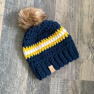 May include: A navy blue crochet beanie with a horizontal stripe of yellow and white. The hat has a faux fur pom-pom on top and a small tag that reads "Stitch Fiesta".