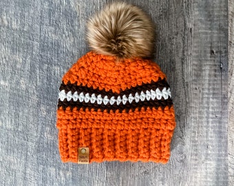 May include: An orange and brown crocheted beanie with a faux fur pom-pom. The hat features a brown and white striped design and a small tag that reads "Stitch Fiesta". The beanie is a warm and stylish accessory.