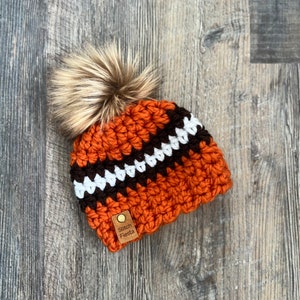 May include: Orange, brown, and white crocheted beanie with a faux fur pom pom. The beanie has a brown leather tag that says "Stitch Fiesta".
