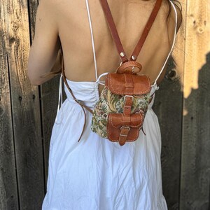 May include: A small backpack featuring a floral tapestry fabric body with brown leather trim, a top flap with a buckle closure, and a front pocket with a buckle. The bag includes thin brown leather shoulder straps and a top carry handle.
